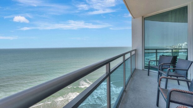 Un balcon avec vue sur l’océan, une balustrade en verre et des fauteuils en osier.