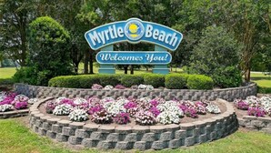 Een bord met 'Myrtle Beach' en 'Welkom!' erop, omringd door bloembedden.