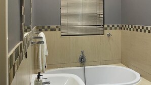 Un baño con bañera, lavamanos, espejo y ventana con persianas.