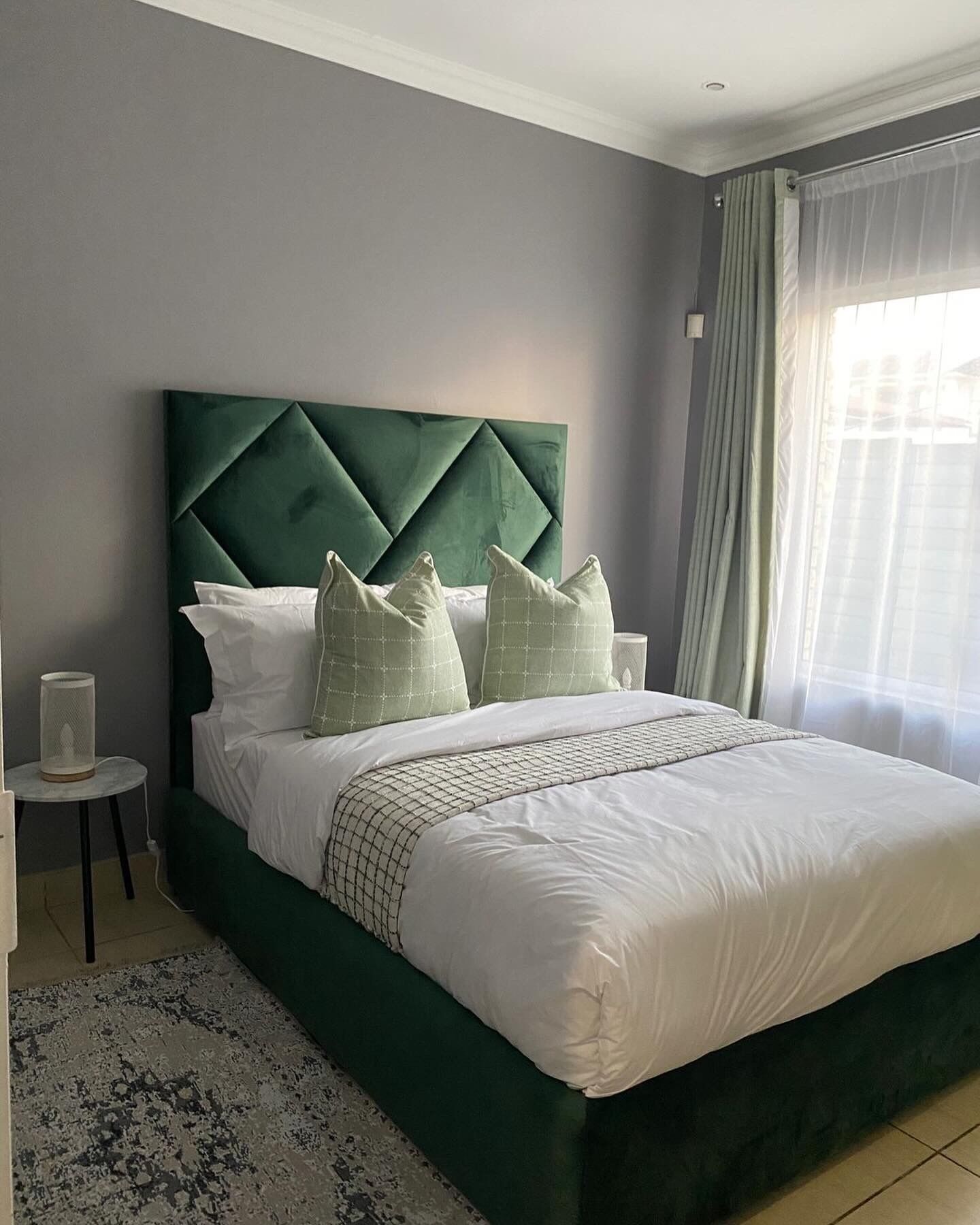 Una cama bien hecha con cabecera tapizada en verde, ropa de cama blanca y almohadas decorativas. Hay una mesita de noche con una lámpara y un objeto decorativo junto a la cama. La habitación tiene una pared gris y una ventana con cortinas de encaje.