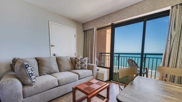 A living room with a sofa, a wooden coffee table, a dining table, and a view of the ocean through a balcony. - Studio Sand Castle South,Direct Ocean View, Pools! (Myrtle Beach)