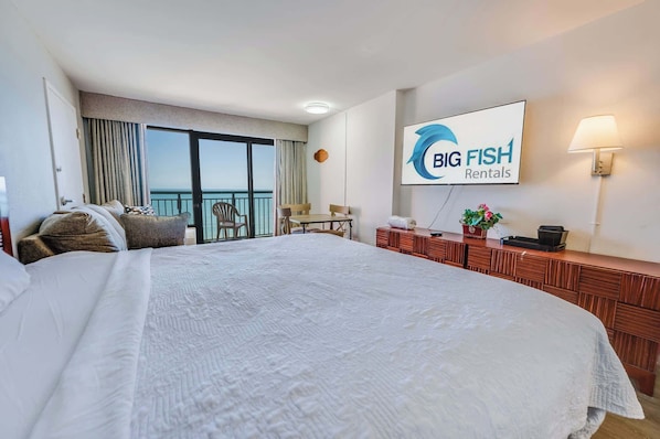 A hotel room with a large bed, a desk, a chair, a balcony with a view, and a sign for Big Fish Rentals.