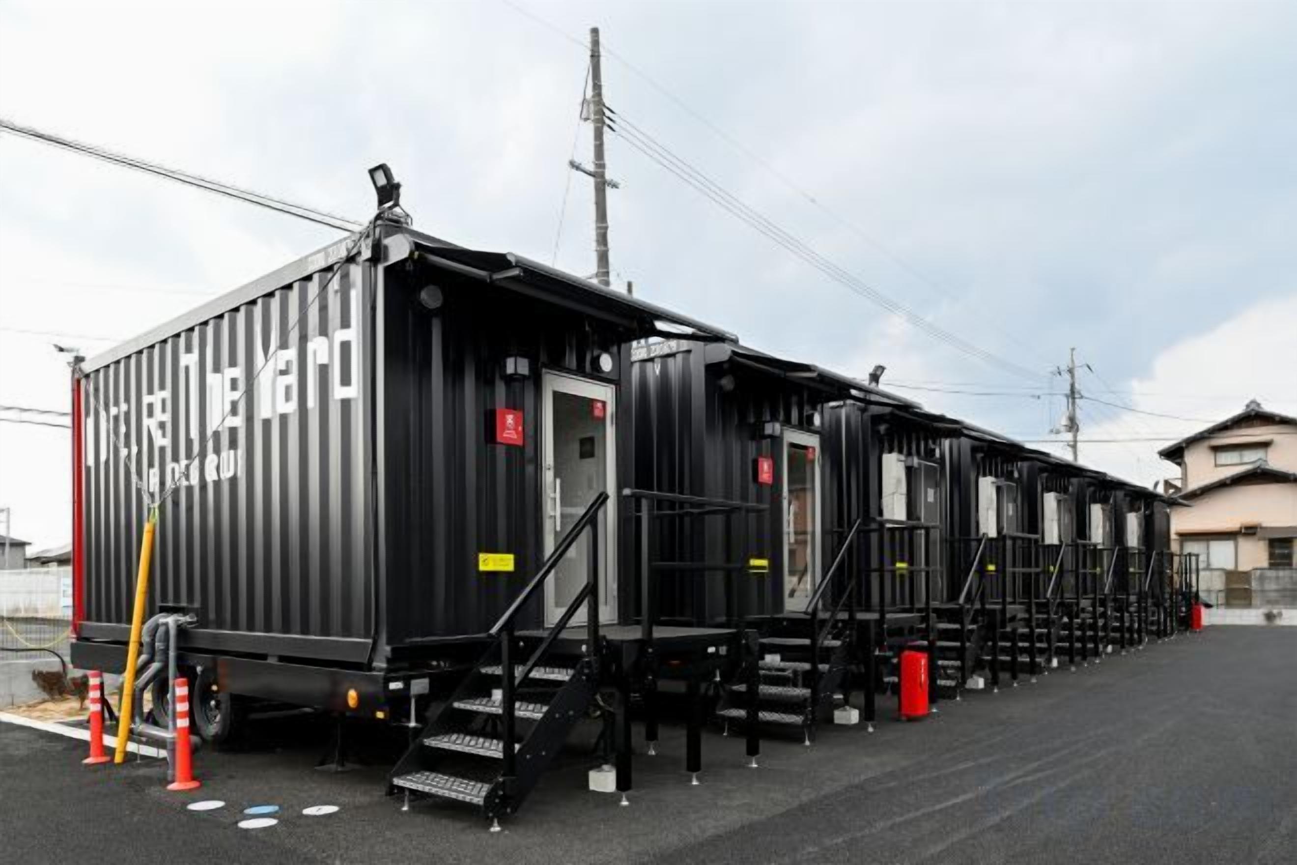 A row of black train carriages with external stairs and a small door, parked on a paved surface with a building in the background.