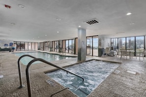 An indoor swimming pool with a hot tub, surrounded by glass walls and outdoor seating.