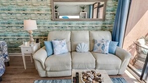 A living room with a beige sofa, a small side table with a lamp, a mirror above the sofa, and a view of the outdoors through a window. - New Direct 1BR Oceanfront Sandcastle South, Pools! (Myrtle Beach)