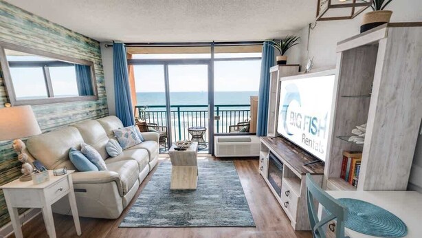 A modern living room with a sofa, coffee table, and TV, overlooking a balcony with a view of the sea. - New Direct 1BR Oceanfront Sandcastle South, Pools! (Myrtle Beach)