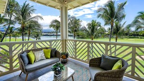 A covered outdoor area with wicker furniture, a glass coffee table, and a view of a golf course and palm trees. - Renovated Penthouse Lake & Golf Course Views AC Near Bay - Vrkcv (Waikoloa)