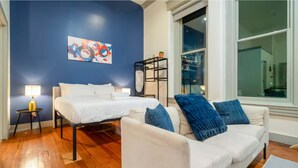 A room with a bed, a white sofa, a small table with a lamp, a window, and a painting on the wall. - Near Hospitals & DT  ~  Vibey+Parking+W/D+Churchill (Louisville)