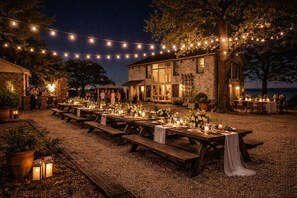 An outdoor dining area with string lights, wooden tables, and benches, set against a stone building with a view of the sea.