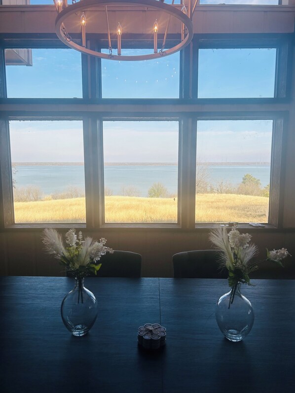 A room with three large windows offering a view of a field and a body of water. There are two vases with flowers on a table.
