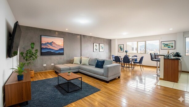A modern living room with a grey sofa, a coffee table, and a TV mounted on the wall. There are framed pictures on the wall and a city view through large windows. - Gran Esmeralda Space View and Style (Cuenca)