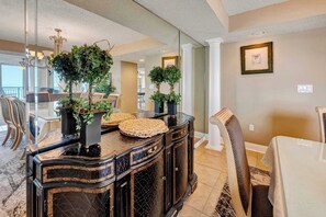 A dining area with a glass-top table, black marble console, potted plants, and a chandelier.