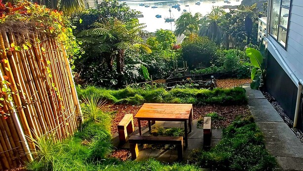 Een tuin met een houten bankje en een bamboe hek, uitkijkend op een jachthaven met boten.