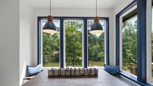 Ein Essbereich mit einem Holztisch, roten Stühlen und einer Bank mit blauen Kissen. Drei große Fenster mit blauen Rahmen bieten Blick auf grüne Vegetation.