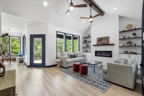 A spacious living room with a large window, a grey sofa, a red ottoman, and a TV mounted on the wall. - Serene Lakeside Retreat, Family and Pet Friendly! (Point Blank)