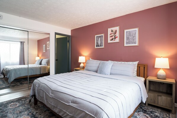 A bedroom with a large bed, two bedside tables with lamps, a mirror, and framed botanical artwork on the wall.