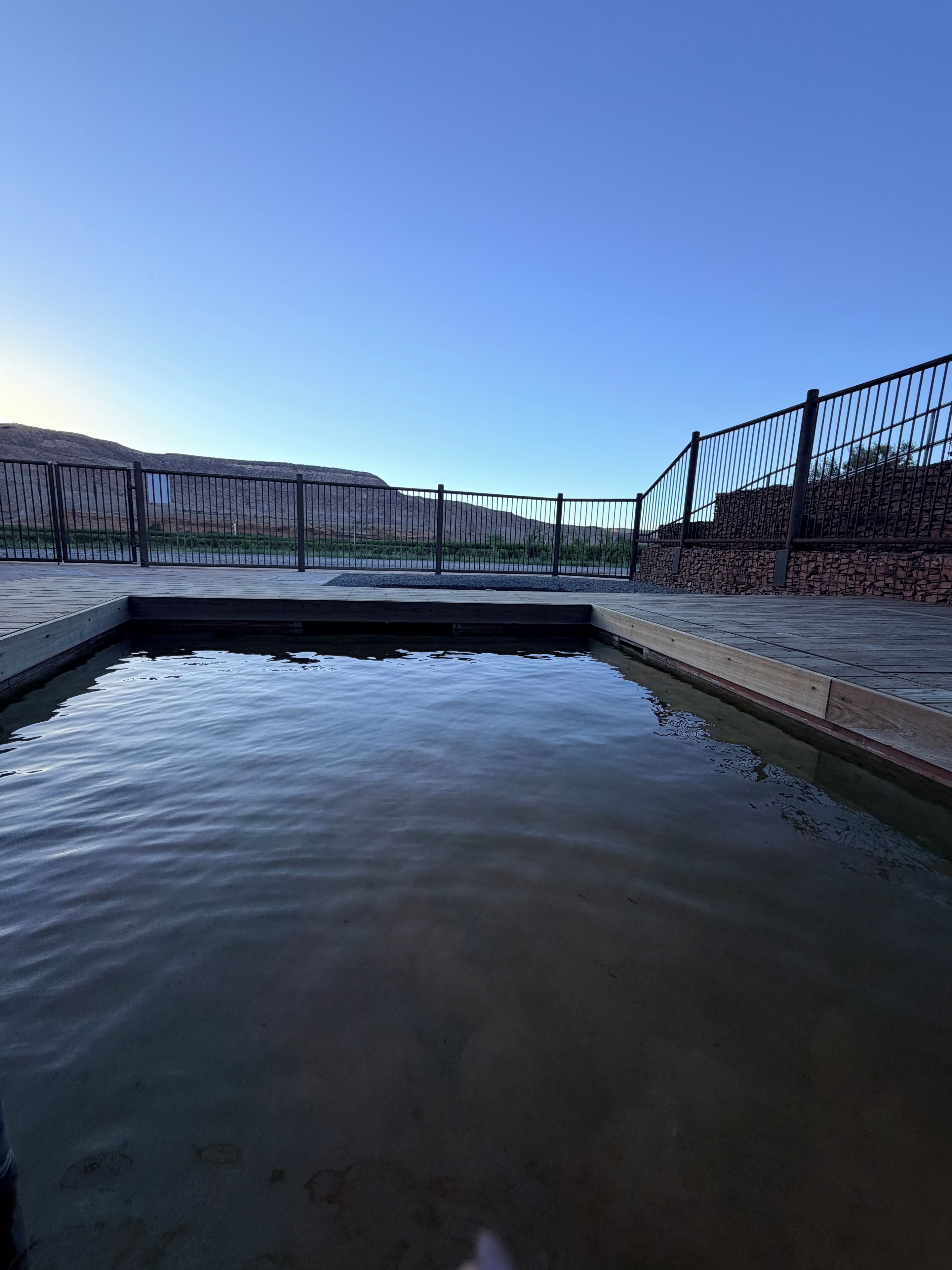 A clear view of a swimming pool with a wooden deck and metal railing, surrounded by a stone wall and a clear sky.