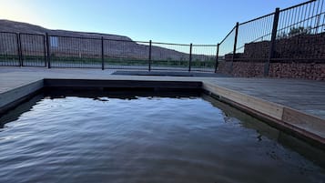 A clear view of a swimming pool with a wooden deck and metal railing, surrounded by a stone wall and a clear sky.