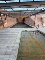 Una piscina con plataforma de madera y un área techada con techo de metal.