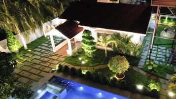 A well-lit residential property with a swimming pool, landscaped garden, and a covered patio area.