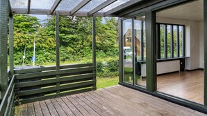 Une terrasse en bois avec une structure vitrée, un banc et une vue sur un paysage verdoyant.