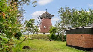 Un moulin à vent au design traditionnel, entouré d’une zone herbeuse avec des arbres et un petit bâtiment en briques.