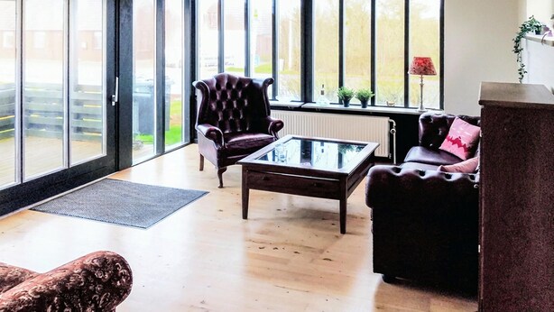 Un salon spacieux, avec un fauteuil en cuir, une table basse en verre et un canapé. De grandes fenêtres offrent une vue sur l’extérieur.