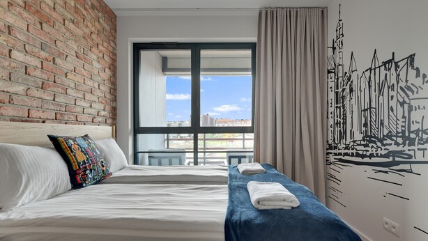 Une chambre d’hôtel avec un mur en briques, une grande fenêtre donnant sur un beau paysage, un lit avec du linge de lit blanc et une serviette bleue posée sur une table d’appoint.