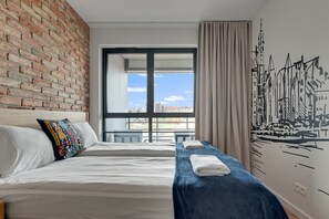 Une chambre d’hôtel avec un mur en briques, une grande fenêtre donnant sur un beau paysage, un lit avec du linge de lit blanc et une serviette bleue posée sur une table d’appoint.
