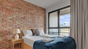 Une chambre avec un mur en briques, une grande fenêtre avec des rideaux, une table de chevet en bois, un lit recouvert d’une couverture bleue et une lampe blanche.