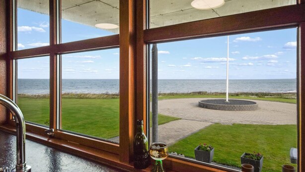 Blick aus einem Fenster auf eine Wiese mit einer runden Steinstruktur und einer Fahnenmast, im Hintergrund das Meer.