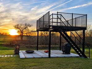 A metal structure with a staircase, a fire pit, and a grill set up in an open field with a sunset in the background.