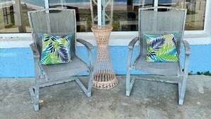 Two wicker chairs with colorful cushions, a woven table, and a view of the beach through large windows.