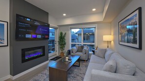 A modern living room with a flat-screen TV mounted on the wall, a fireplace, a grey sofa, a coffee table, and a large window offering a view of the outside.