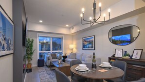 A modern living room with a dining area, a flat-screen TV, a chandelier, and a large window with a view.