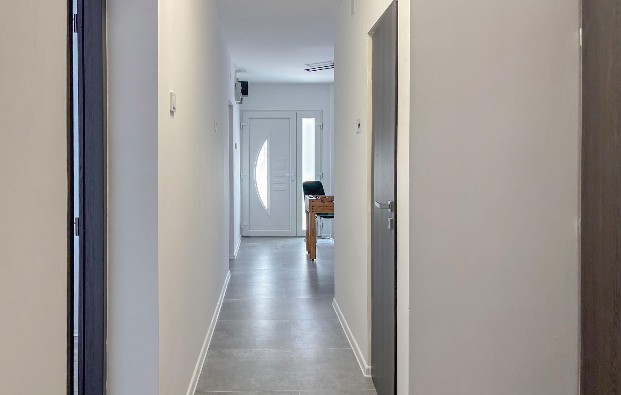 A narrow hallway with a door at the end, a small table with a chair, and a window with blinds.