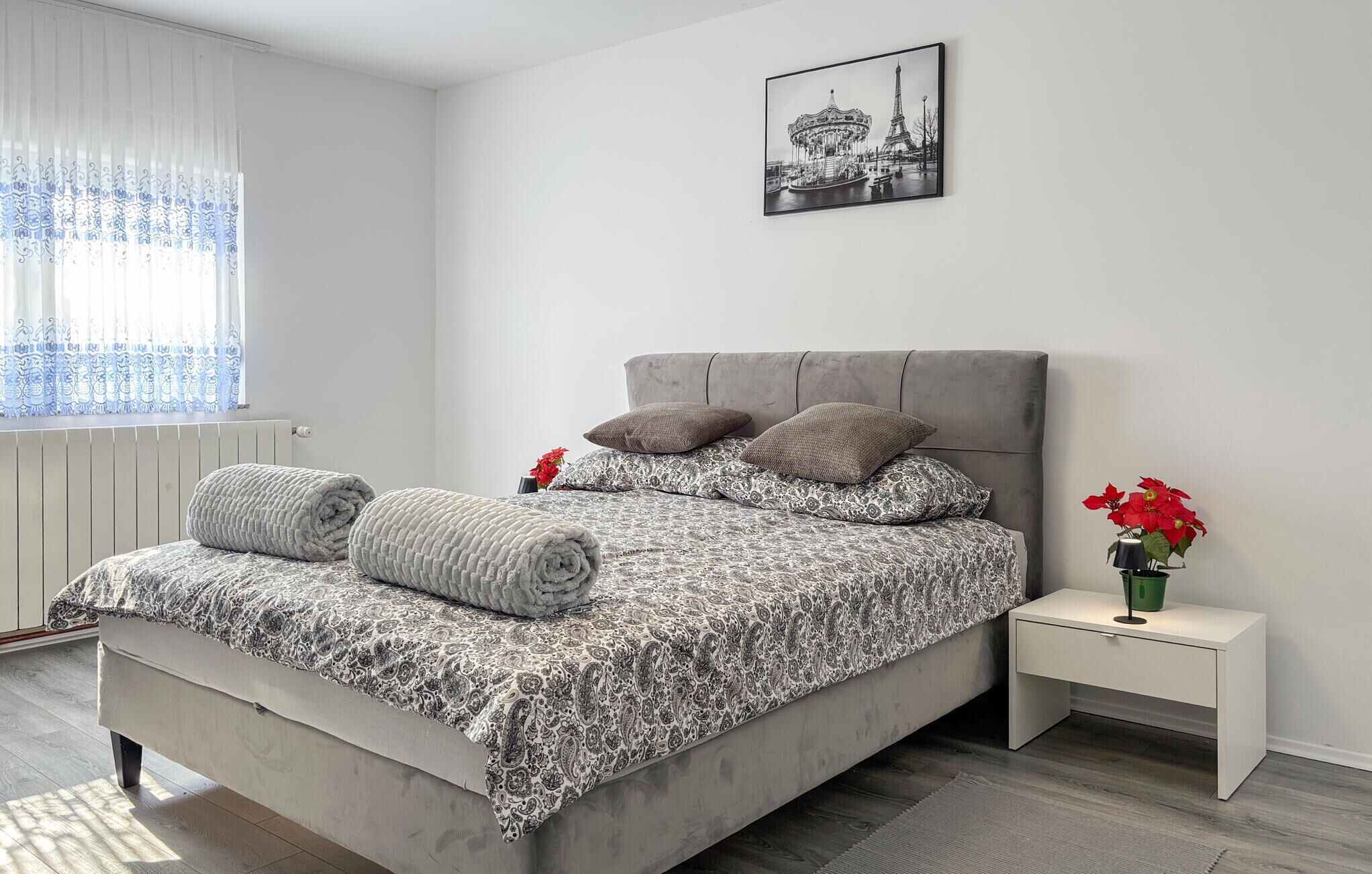 A bedroom with a bed, pillows, a nightstand, a vase with flowers, and a picture on the wall.