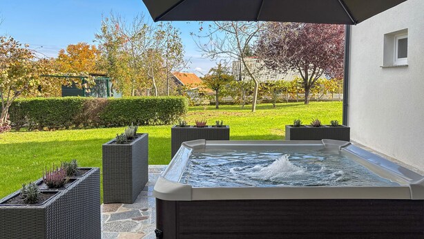 An outdoor hot tub area with a stone-tiled floor, surrounded by wicker planters with plants, and a view of a well-maintained lawn and trees. - Stunning home in Salopeki Modruski with WiFi (Salopeki Modruski)