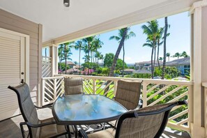 Een overdekt buitengebied met een glazen tafel en stoelen, een witte balustrade en uitzicht op palmbomen en huizen.