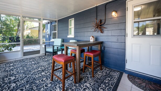 A covered porch with a table and four red stools, a decorative wall piece, and a white door. - Charming Remodeled Cottage | Walk to Park Ave (Winter Park)