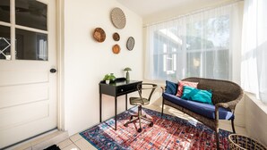 A room with a door, a desk with a lamp, a wicker chair with cushions, a patterned rug, and wall decorations. - Charming Remodeled Cottage | Walk to Park Ave (Winter Park)
