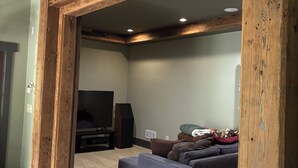Un salón amplio con techo alto y vigas de madera a la vista, un televisor de pantalla plana grande, un sofá de color oscuro y un perro blanco pequeño.
