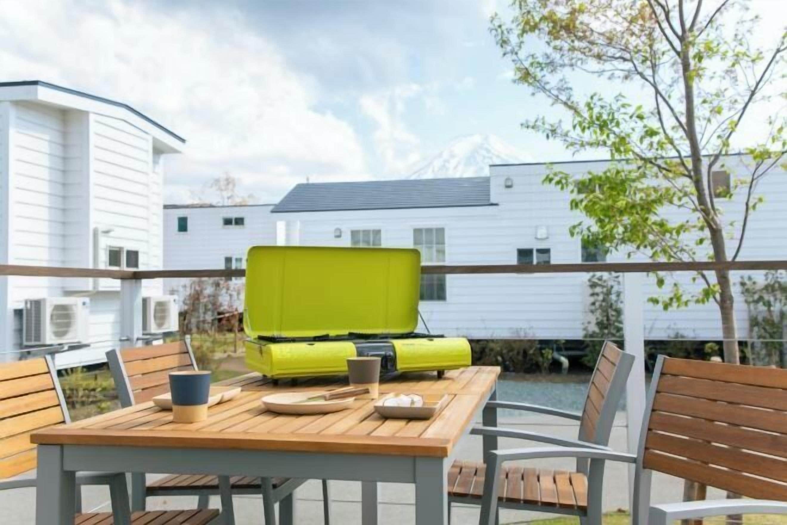 An outdoor seating area with a wooden table and metal chairs, a bright yellow suitcase, and a view of white buildings in the background.