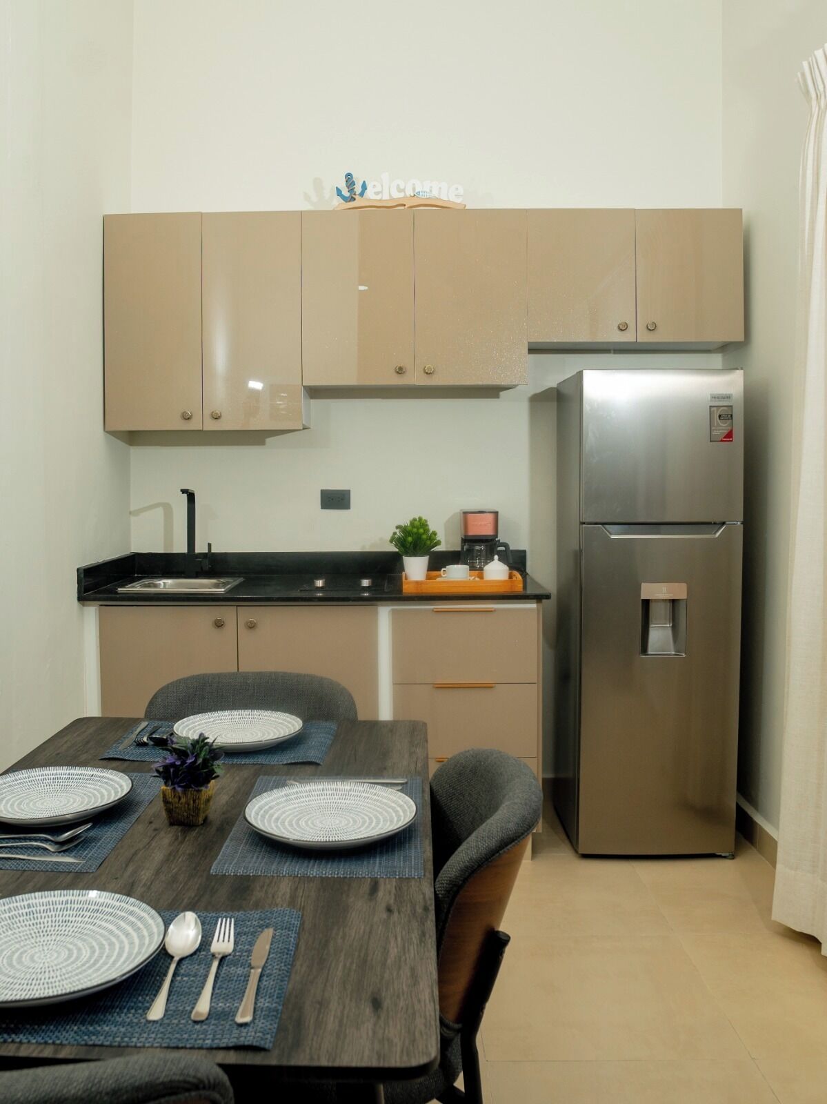 Una cucina moderna con mobili color beige, un frigorifero in acciaio inossidabile e un'area pranzo con un tavolo di legno apparecchiato per quattro persone.