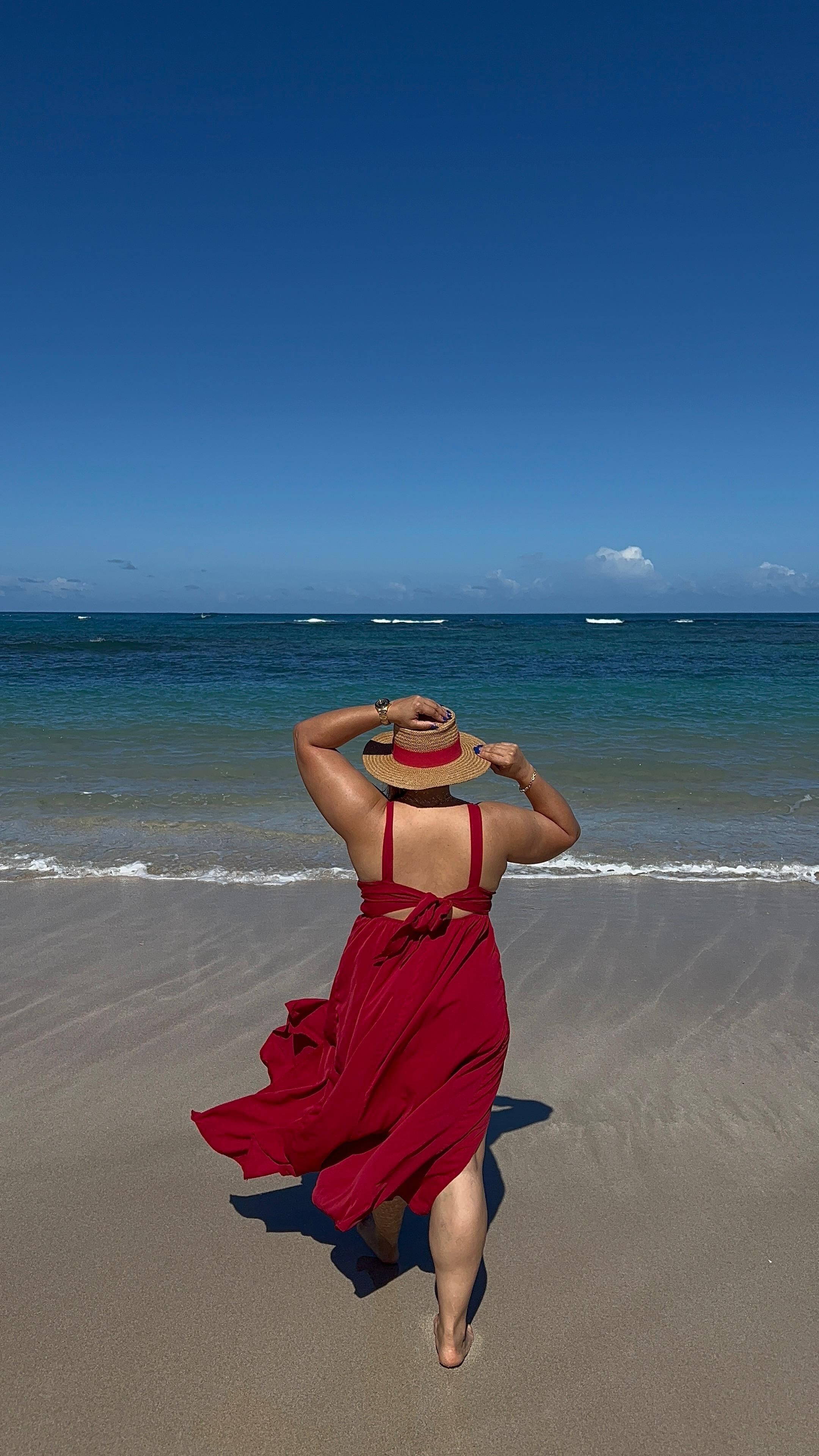 A person in a red dress walking on a sandy beach with the ocean in the background.