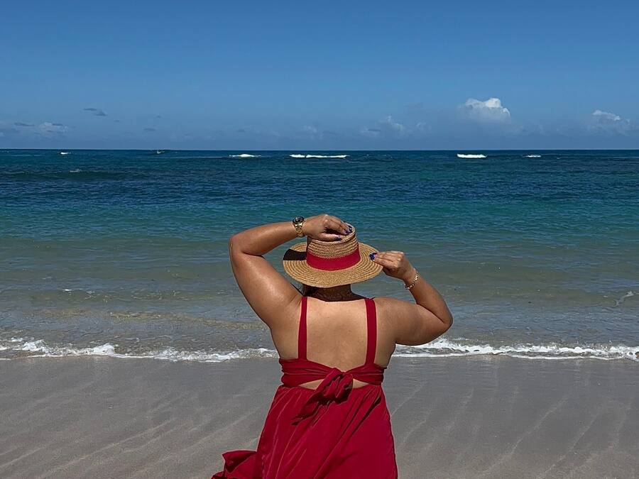 A person in a red dress walking on a sandy beach with the ocean in the background.