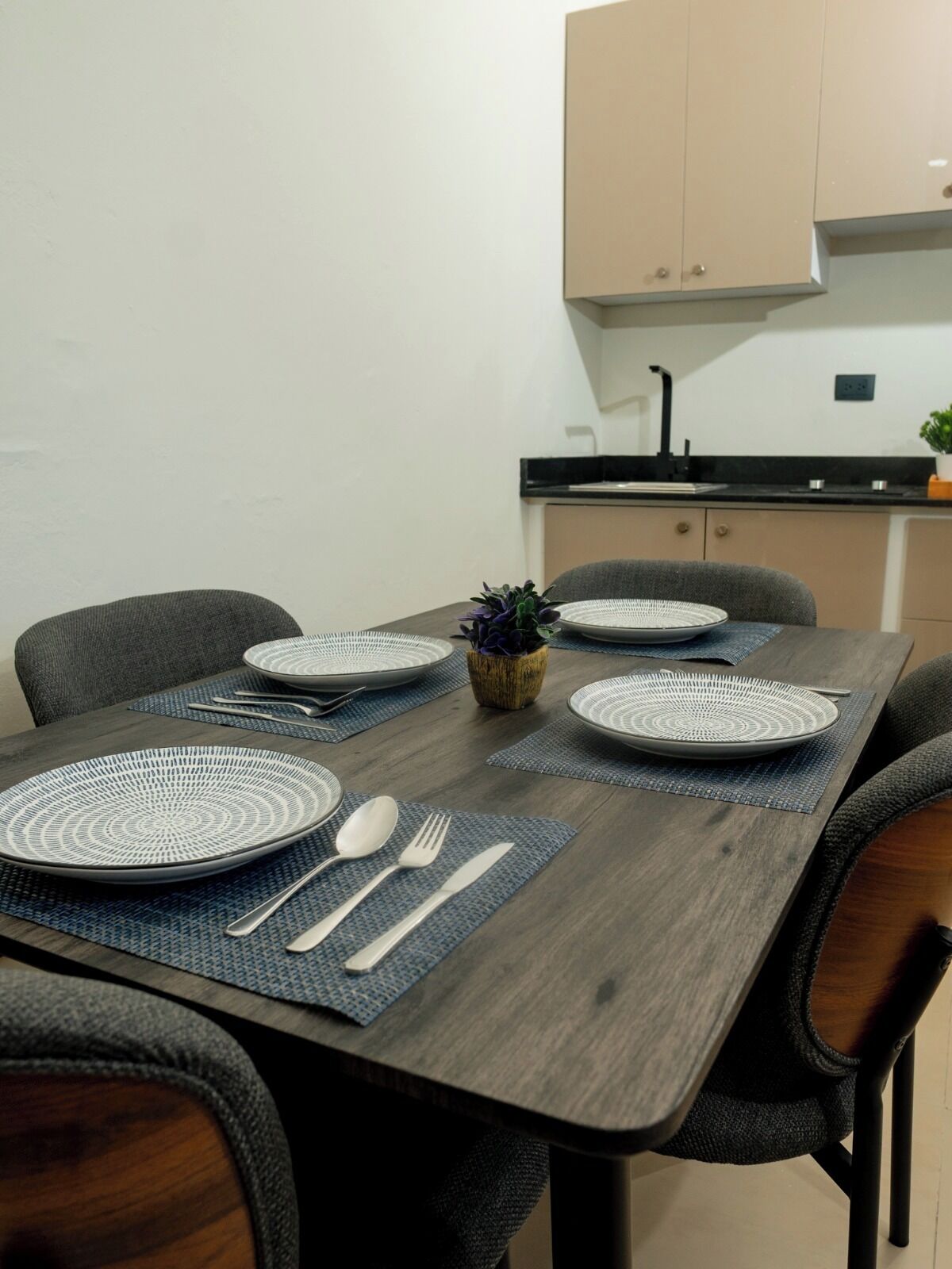 Una mesa de comedor dispuesta con platos, cubiertos y posavasos en una cocina moderna con armarios y un lavamanos.