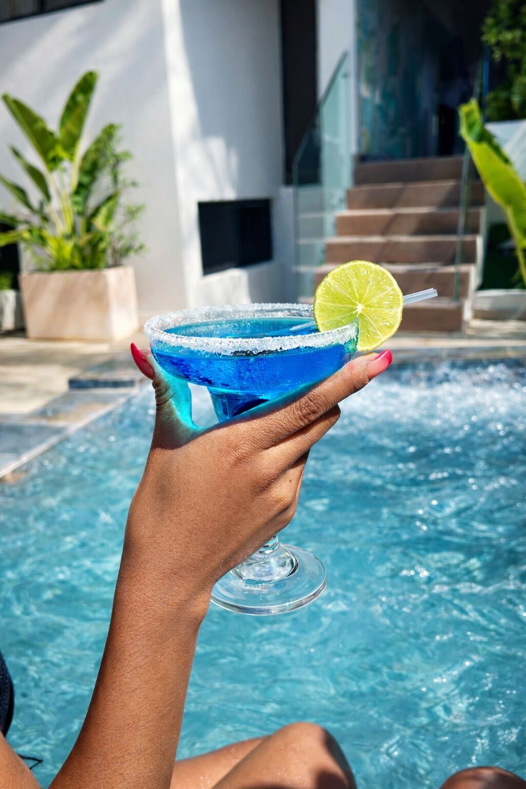 Een hand die een blauwe cocktail met een limoenpartje vasthoudt, in de buurt van een zwembad.