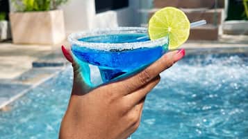 Een hand die een blauwe cocktail met een limoenpartje vasthoudt, in de buurt van een zwembad.