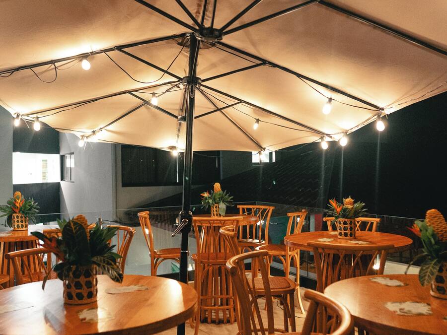 Un espacio exterior para comer con varias mesas y sillas de madera, cubierto por grandes sombrillas y luces colgantes.